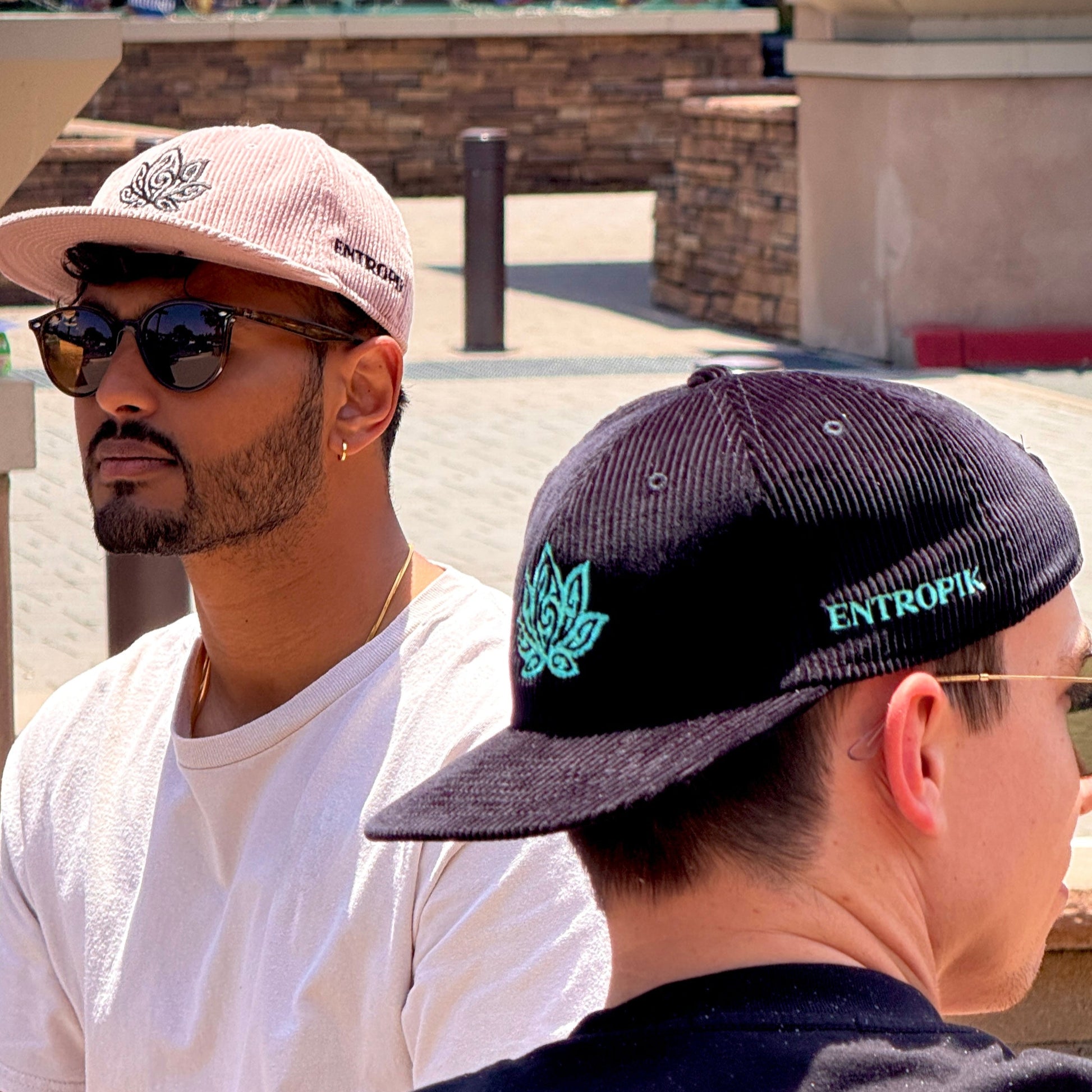 Two men, one wearing a white t-shirt and pink cap, the other wearing a black cap with a logo, standing outdoors.